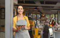 Woman in an apron holding a tablet, outside of a storefront.