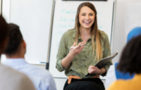 Young woman holding a notebook and smiling at her colleagues, in front of a whiteboard.