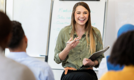 Young woman holding a notebook and smiling at her colleagues, in front of a whiteboard.